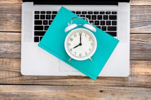 Laptop, book and white alarm clock on wooden background, top view.
